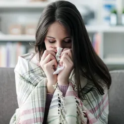 A woman sitting on a couch holding Kleenex up to her nose